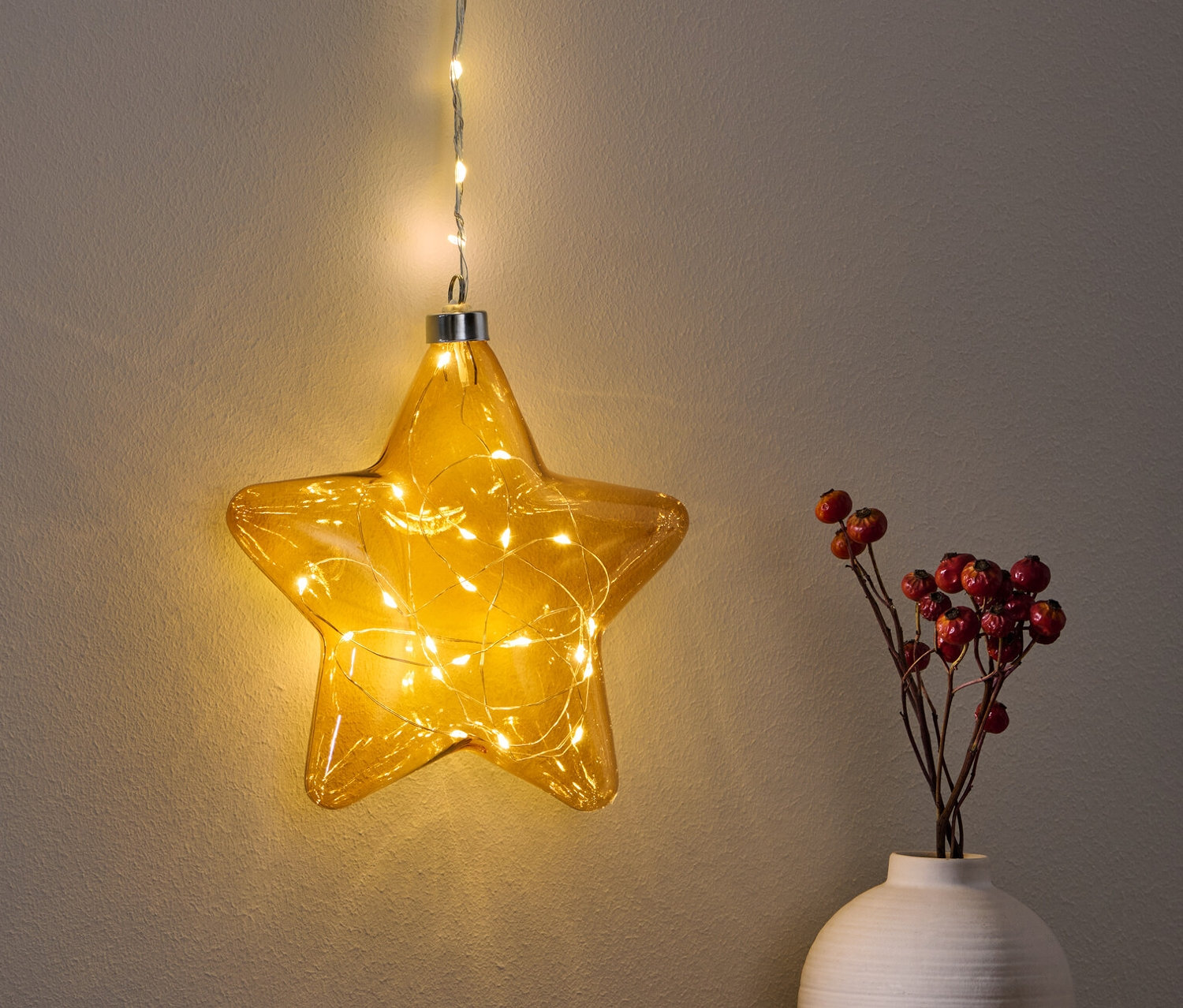 Ein hängender LED-Stern leuchtet über einem Tisch neben einer Vase mit roten Beeren.