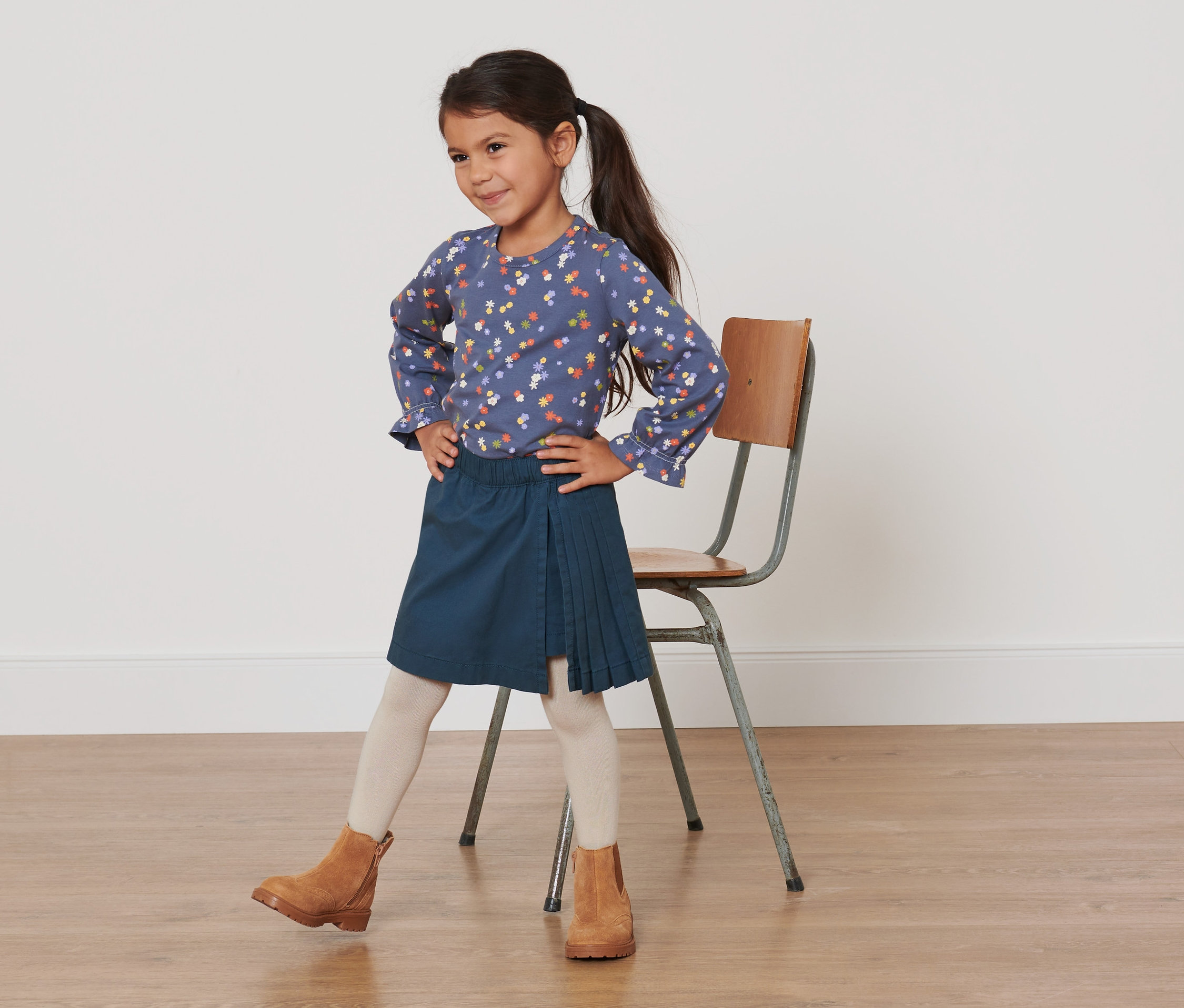 Mädchen posiert in blauer Bluse mit Blumenmuster und dunkelblauem Faltenrock. Sie trägt einen Kleinkinder-Kiltrock.