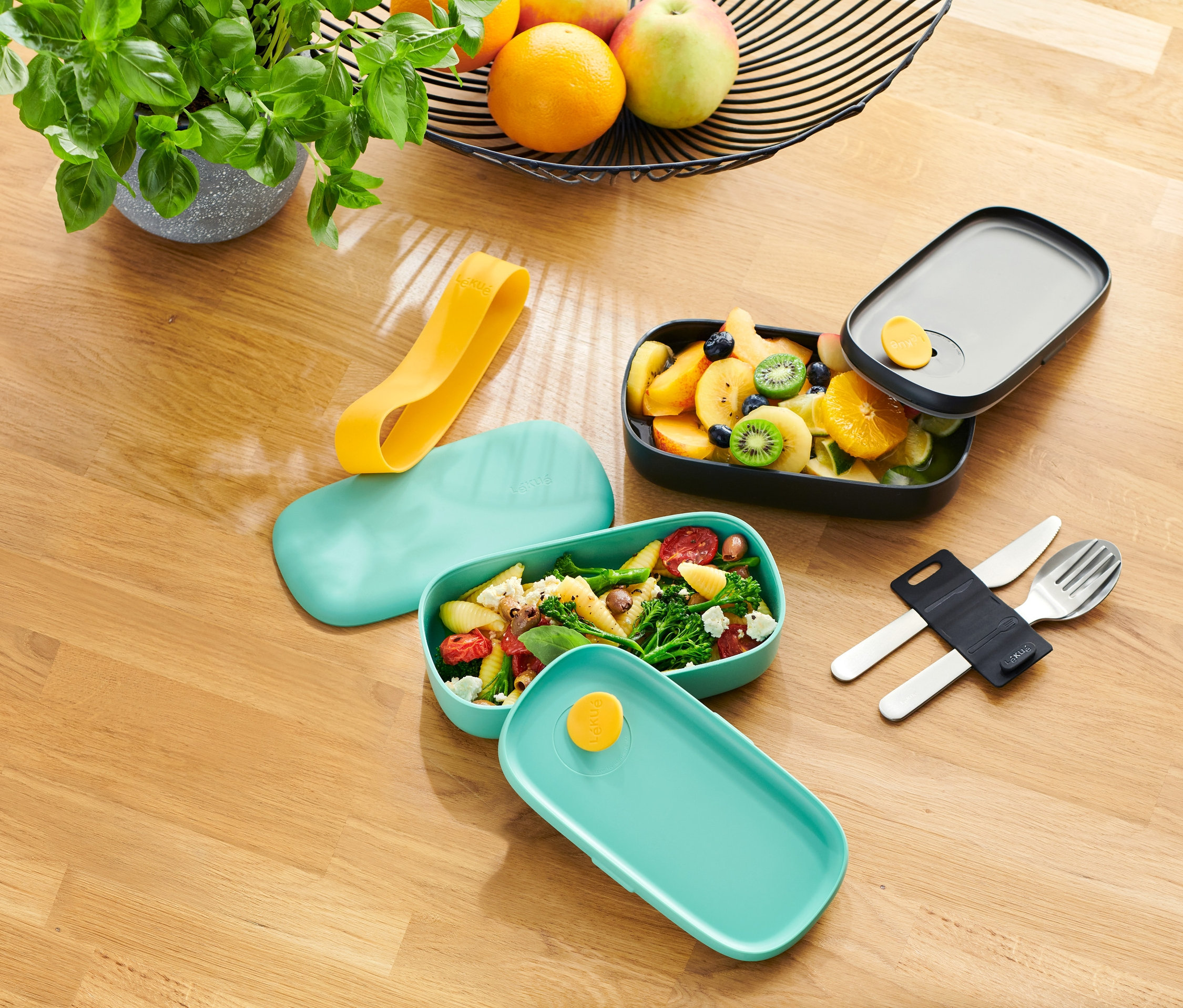 Zwei geöffnete Lunch-Box-Sets mit Besteck stehen auf einem Holztisch. Eine Dose ist mit Obst und die andere mit Gemüse gefüllt.