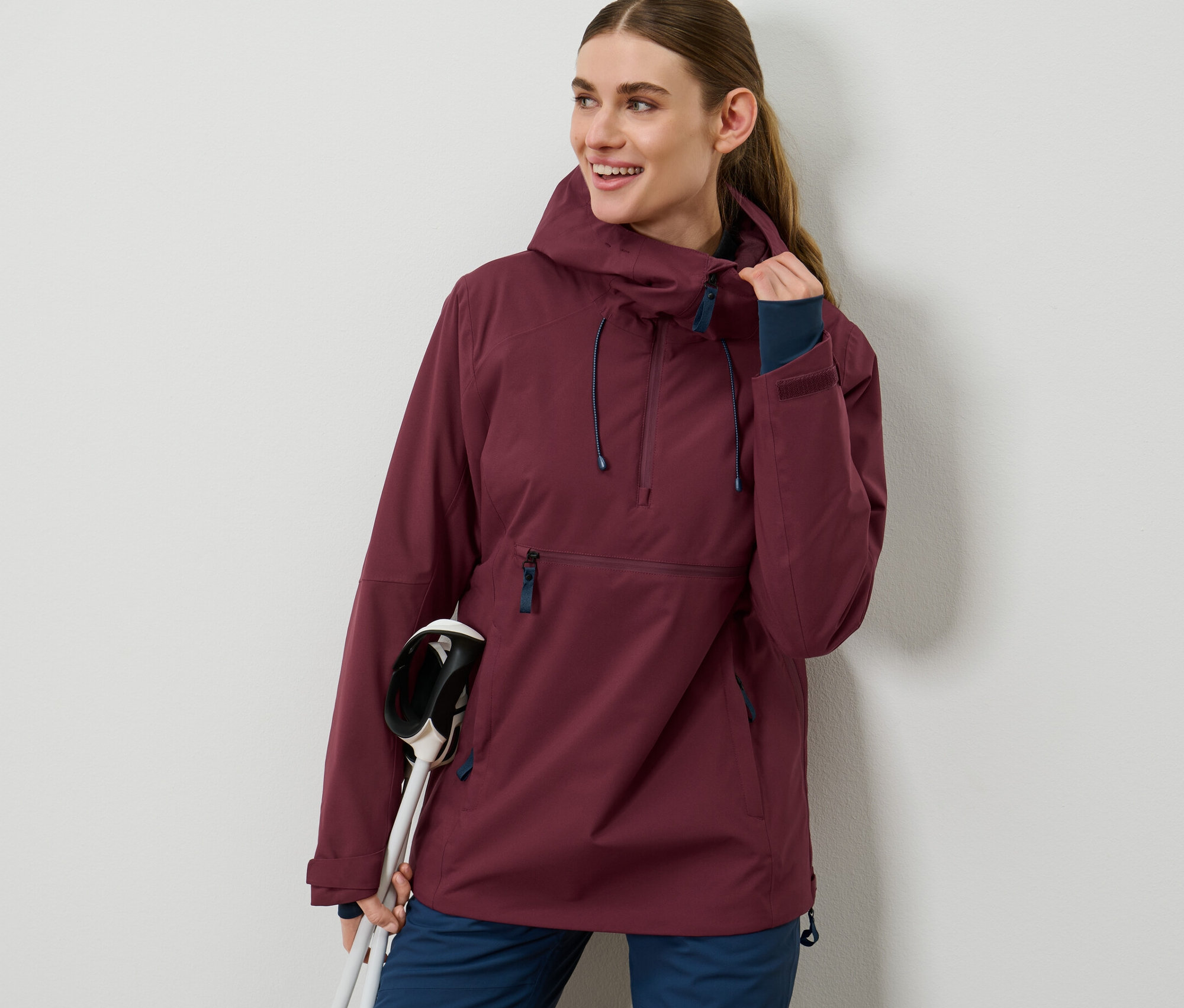 Frau trägt eine bordeauxrote Halfzip-Skijacke und hält Skistöcke.