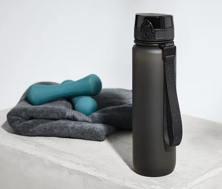 Eine Trinkflasche steht auf einer grauen Oberfläche neben einem Handtuch und Hanteln.
