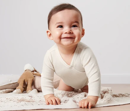 Lächelndes Baby im Baby-Body mit Modal krabbelt auf einer beige Strickdecke neben einem Hasen-Spielzeug.
