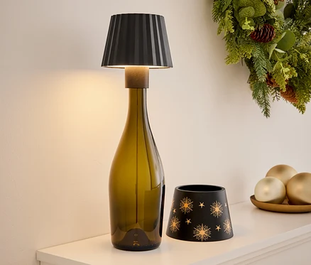 Auf einem weißen Regal stehen eine Lampe aus einer olivgrünen Flasche, ein schwarzer Kerzenhalter mit goldenen Sternen und Dekorationen auf einem Teller. Im Hintergrund befindet sich ein Weihnachtskranz.