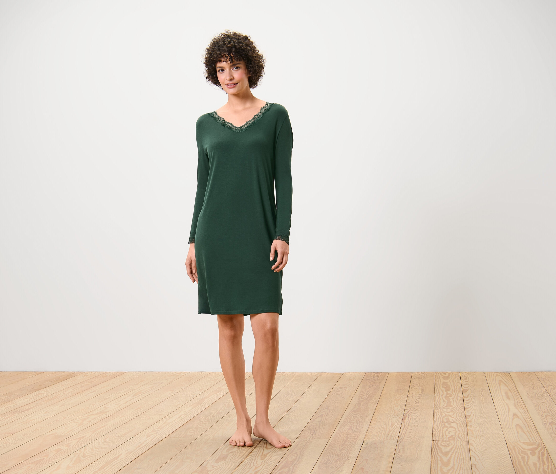 Eine Frau steht barfuß auf einem Holzboden und trägt ein dunkelgrünes Nachthemd mit langen Ärmeln und Spitze am Ausschnitt.