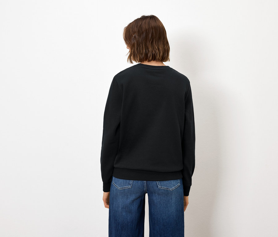 Von hinten ist eine Frau mit braunen Haaren in blauen Jeans und einem schwarzen Pullover zu sehen.