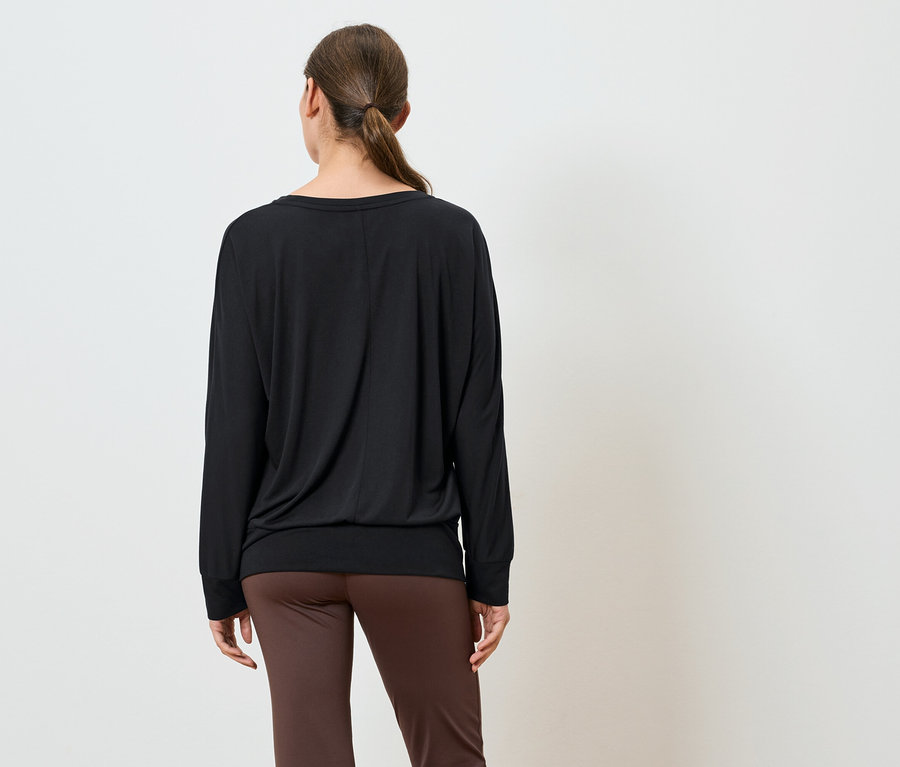 Eine Frau steht mit dem Rücken zum Betrachter und trägt ein schwarzes Langarmshirt und braune Hosen.