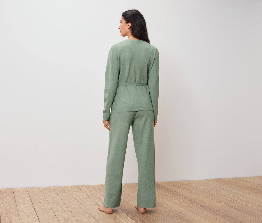 Frau in grünem Pullover und Hose steht mit dem Rücken zum Betrachter auf einem Holzboden.