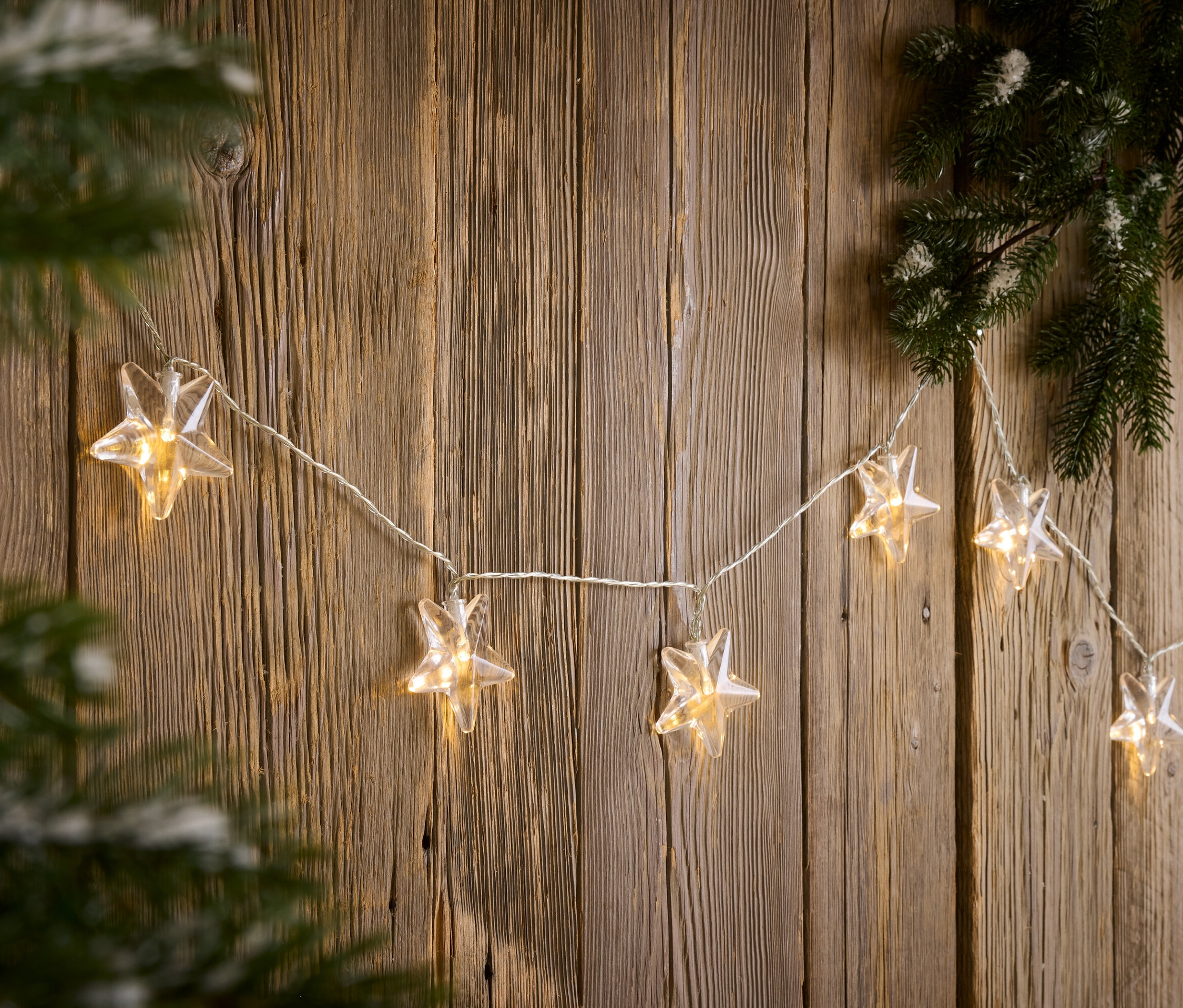 Eine Lichterkette in Sternform hängt an einer Holzwand neben einem Kiefernzweig.