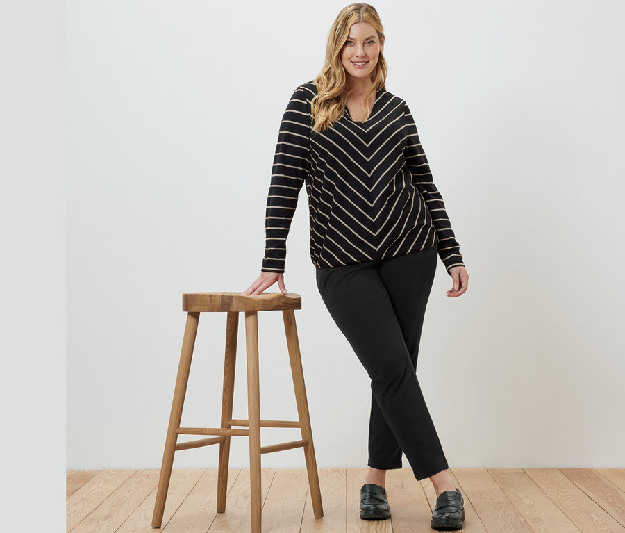 Eine Frau posiert an einem Holzhocker, sie trägt ein gestreiftes Blusenshirt aus Modal-Mix und eine schwarze Joggpants.