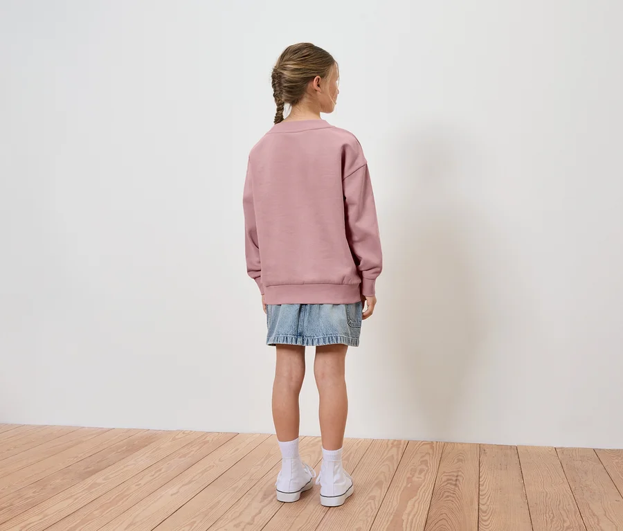 Mädchen mit Zopf trägt einen rosafarbenen Pullover und einen Jeansrock, von hinten gesehen.
