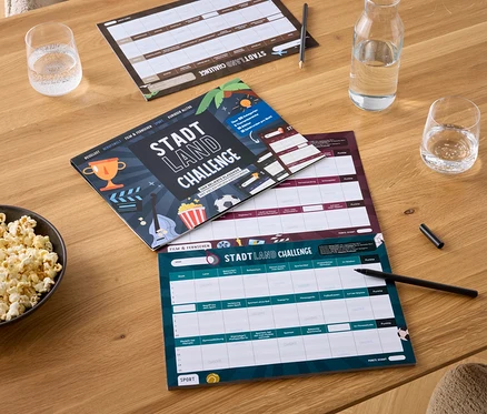 Auf einem Holztisch liegen eine Schüssel Popcorn, zwei Gläser Wasser, eine Wasserflasche und drei Spielbretter Stadt Land Challenge mit Stiften.