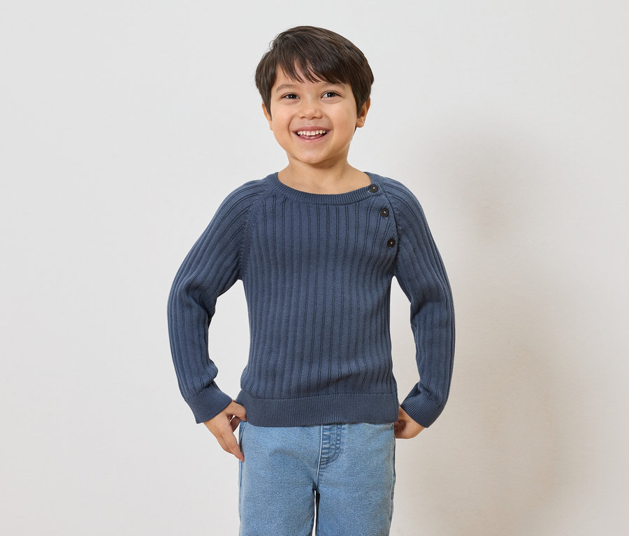 Ein lächelnder Junge mit braunen Haaren trägt einen gerippten, marineblauen Pullover mit Knöpfen und hellblaue Jeans.