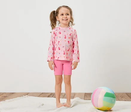 Ein Mädchen mit Zöpfen trägt ein rosa Langarmshirt mit Seepferdchenmuster und pinke Shorts. Ein bunter Ball liegt auf einem weißen Teppich.
