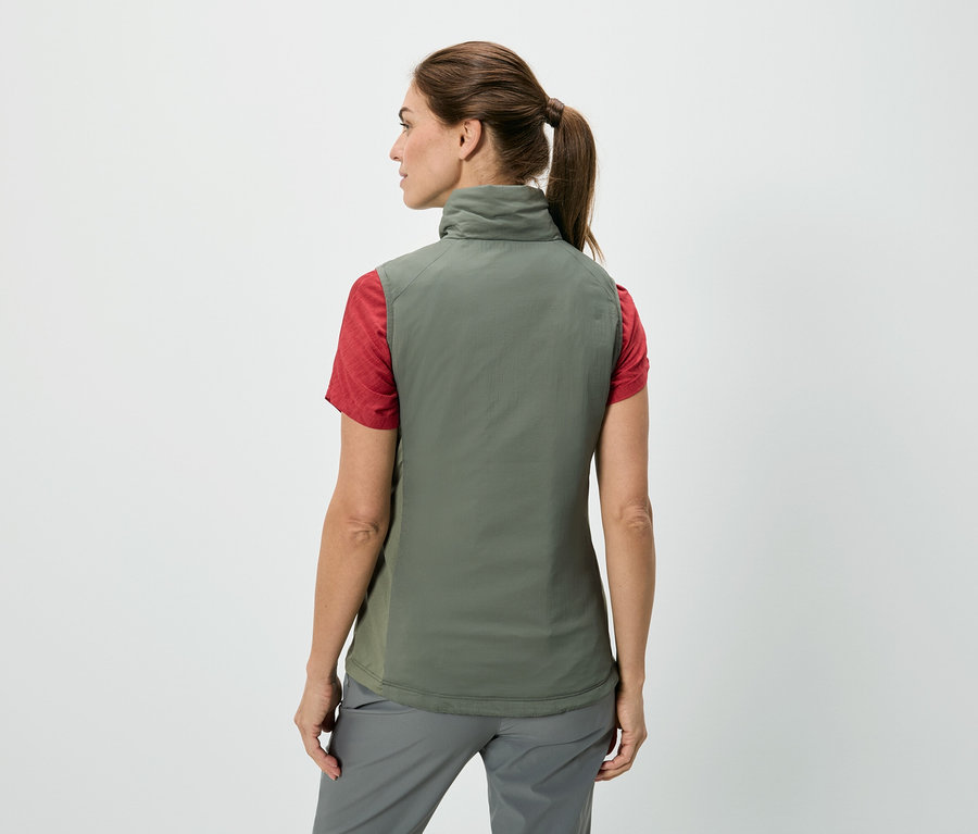 Rückansicht einer Frau mit Pferdeschwanz, die eine olivgrüne Weste über einem roten T-Shirt und grauen Hosen vor einem weißen Hintergrund trägt.