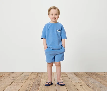 Ein Junge steht vor einer weißen Wand, gekleidet in ein blaues T-Shirt mit Hai-Motiv und blaue Shorts, dazu trägt er blaue Flip-Flops.