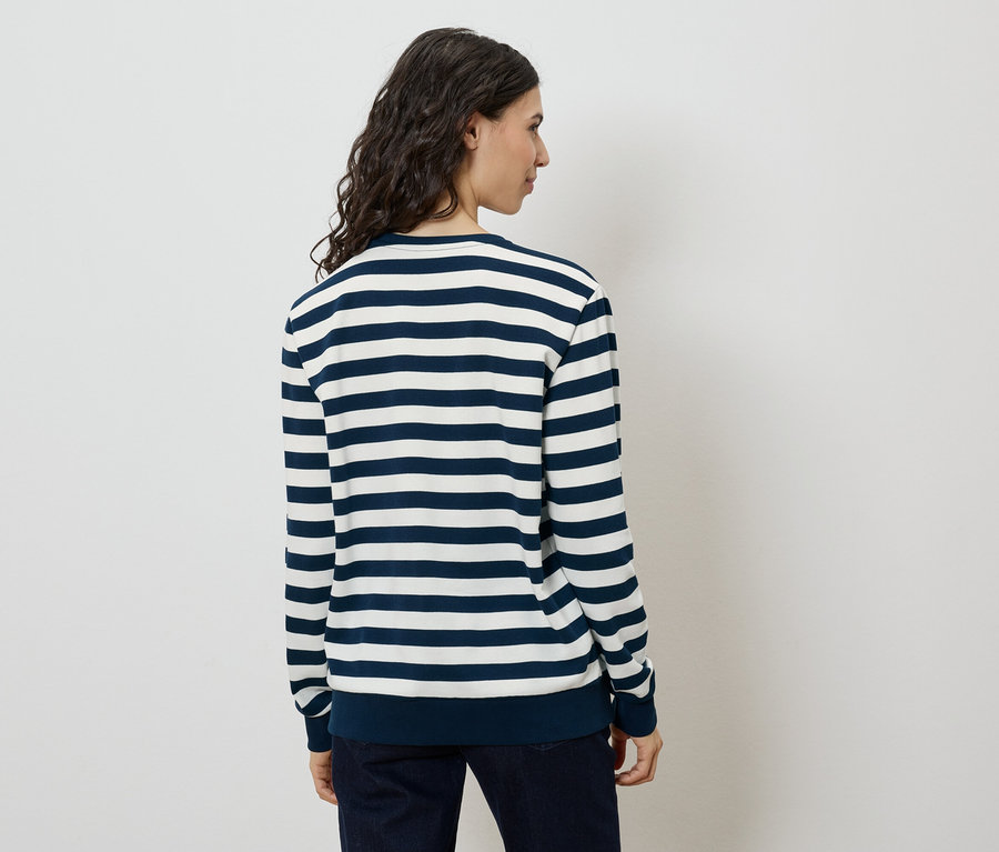 Rückansicht einer Frau mit langen, gewellten, dunklen Haaren, die einen blau-weiß gestreiften Pullover und dunkle Jeans trägt.