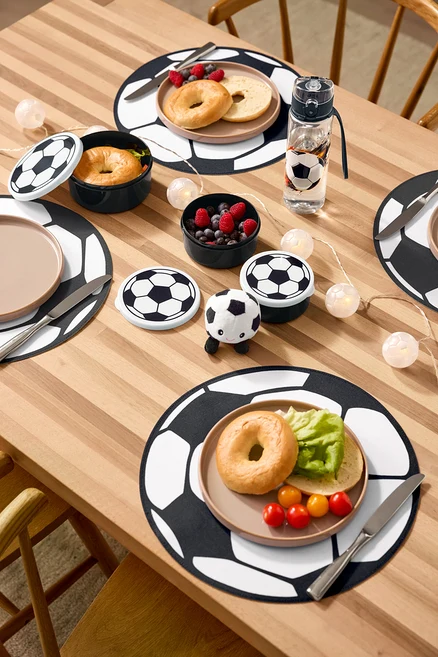 Auf einem Holztisch liegen vier Platzsets, eine Trinkflasche, drei Brotdosen, ein Plüsch-Fußball mit Sound und eine LED-Lichterkette »Fußball«.
