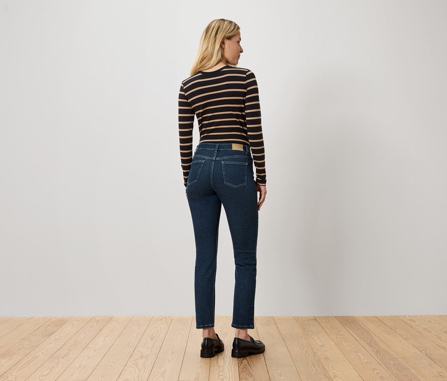 Eine Frau mit blonden Haaren steht mit dem Rücken zur Kamera und trägt ein gestreiftes Langarmshirt, blaue Jeans und schwarze Schuhe.