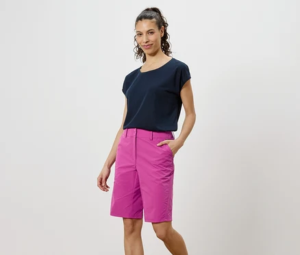 Frau mit dunklem T-Shirt und pinken Shorts steht vor einem weißen Hintergrund.