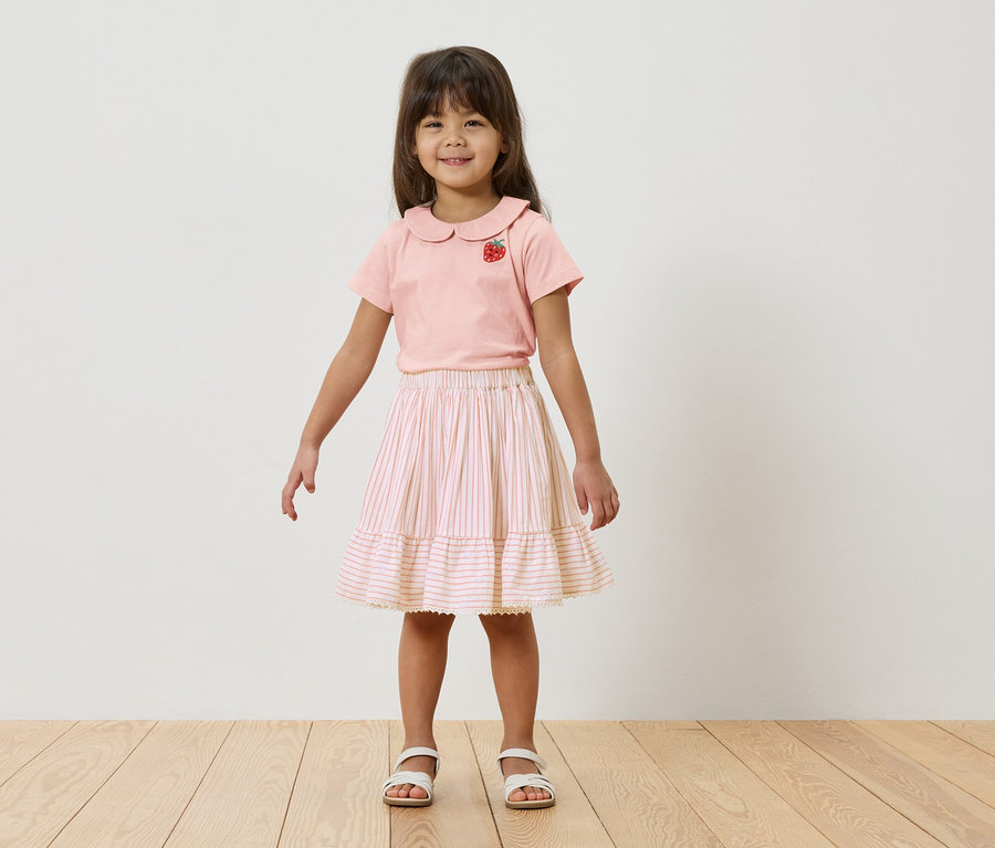 Ein junges Mädchen steht vor einer weißen Wand, sie trägt ein rosa T-Shirt mit Erdbeermotiv und einen gestreiften Rock.