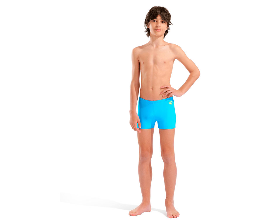 Ein junger Mann steht barfuß vor einem weißen Hintergrund und trägt eine blaue Badehose.