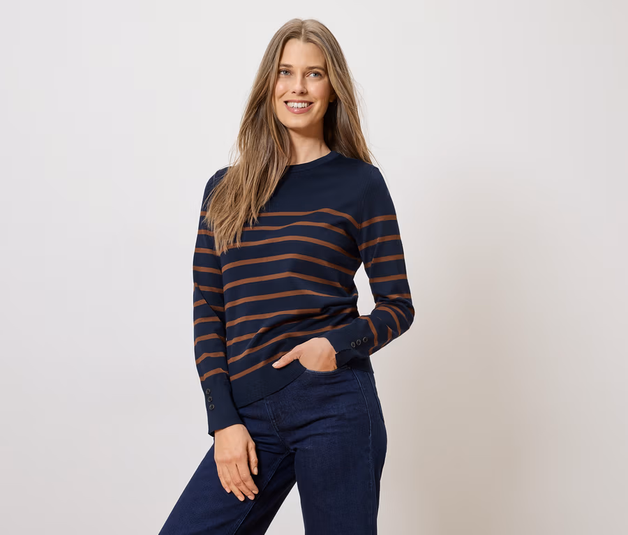 Frau mit einem dunkelblauen, gestreiften Pullover und Jeans, eine Hand in der Hosentasche.