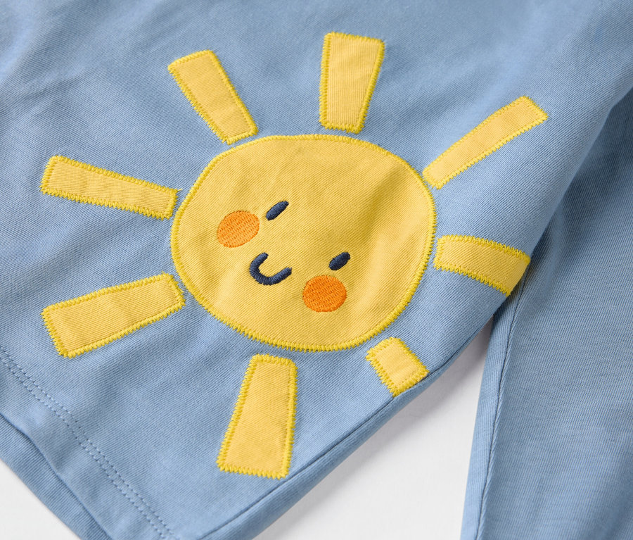 Detailansicht eines hellblauen Kleidungsstücks mit einer aufgenähten gelben Sonne.