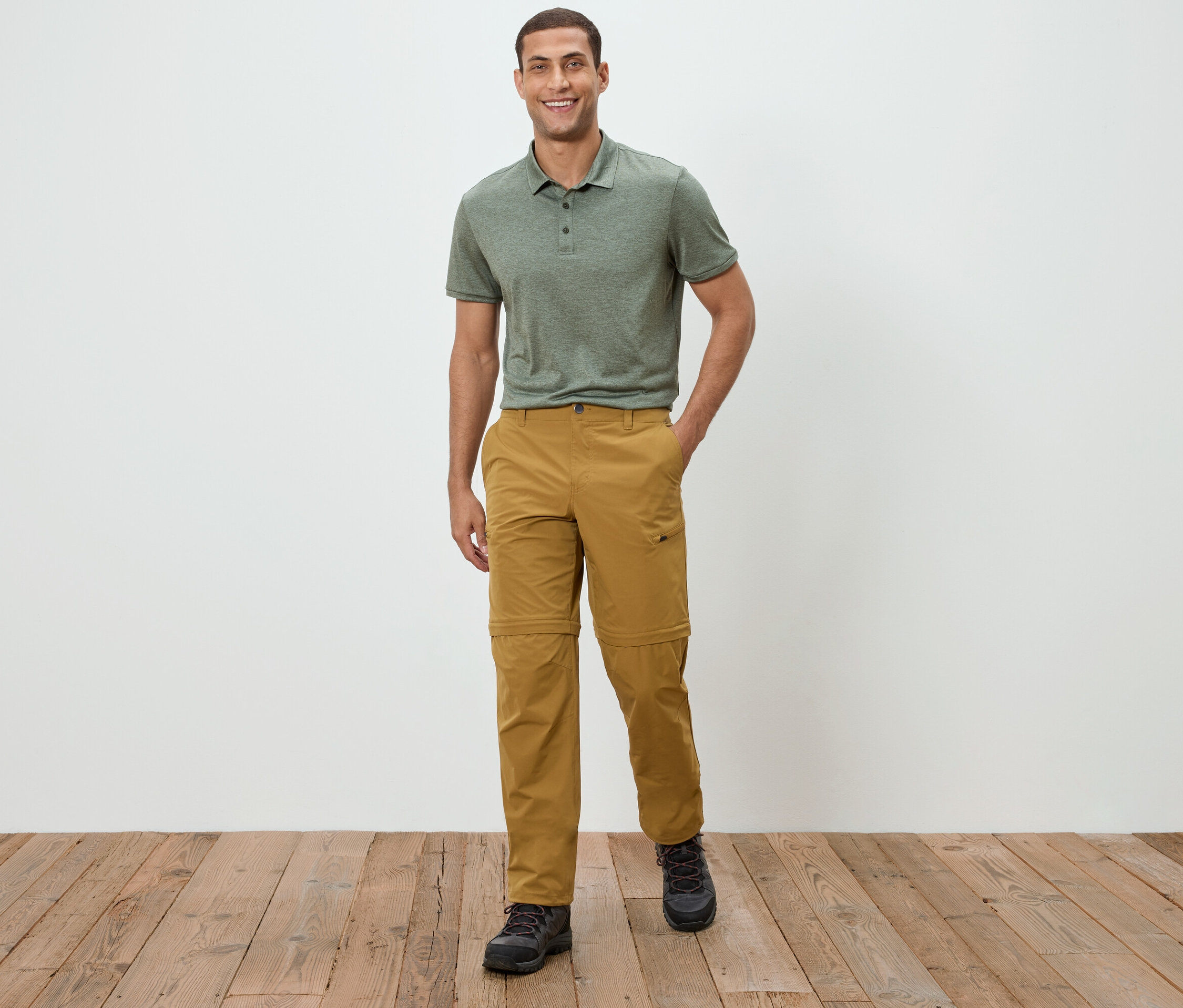 Ein Mann steht vor einer weißen Wand und trägt ein grünes Polohemd und Khakihosen.