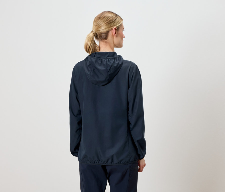 Rückansicht einer Frau mit blonden Haaren, die eine marineblaue Jacke mit Kapuze und marineblaue Hosen trägt.