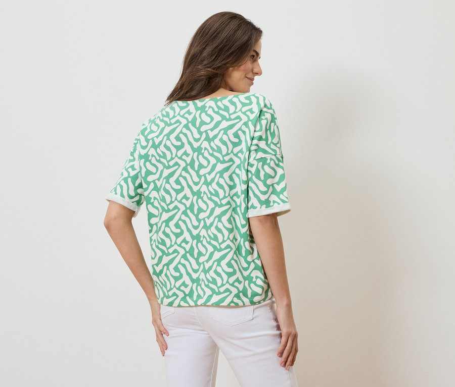 Rückansicht einer Frau mit braunen Haaren, die ein kurzärmeliges, grün-weiß gemustertes Oberteil und weiße Jeans trägt.