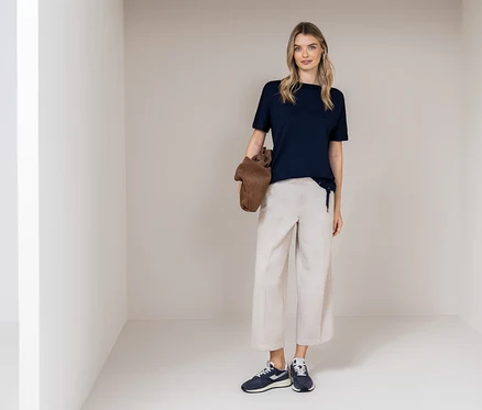 Eine Frau steht in einem Raum und trägt ein dunkelblaues T-Shirt, beigefarbene Hosen und blaue Turnschuhe. Sie hält eine braune Tasche.