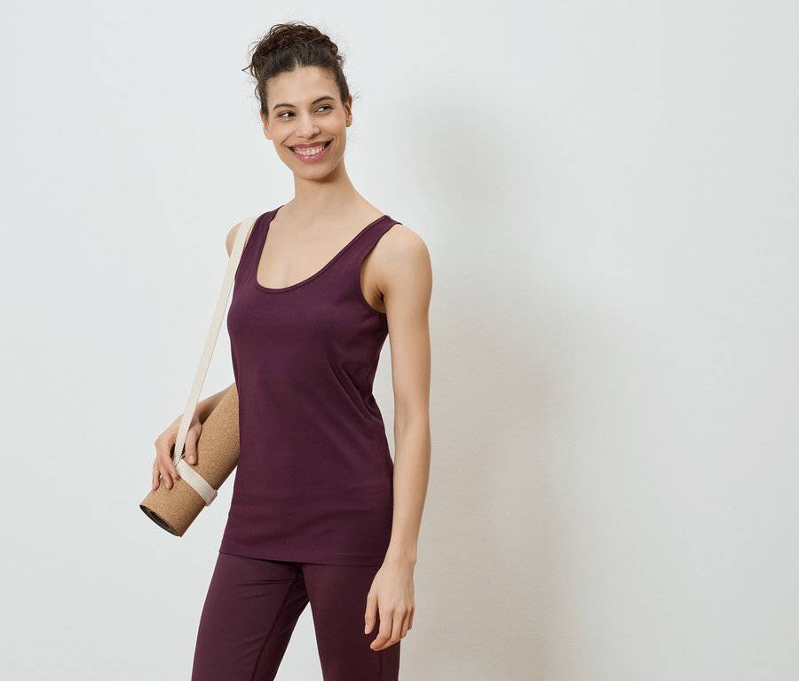 Frau im auberginefarbenen Top und Hose, Yogamatte über der Schulter, lächelt vor einem weißen Hintergrund.