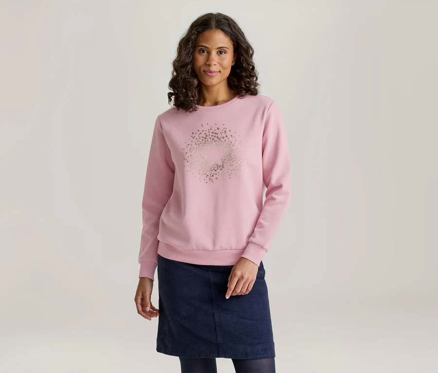 Eine Frau mit dunklem, lockigem Haar lächelt leicht und trägt ein hellrosa Langarm-Sweatshirt mit einem kreisförmigen Sternenmuster auf der Vorderseite, dazu einen dunkelblauen Rock und dunkelblaue Strumpfhosen.