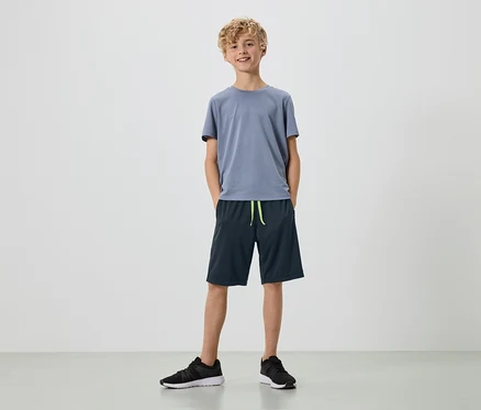 Ein junger Mann mit blonden Locken steht in einem grauen T-Shirt, dunkelblauen Shorts und schwarzen Turnschuhen vor einem weißen Hintergrund.