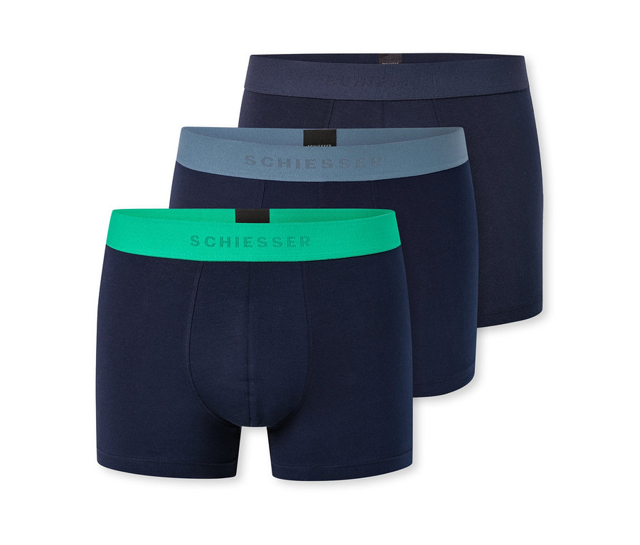 Drei dunkelblaue Herren-Boxershorts sind diagonal gestapelt. Die vorderste hat einen leuchtend grünen Bund, die mittlere einen graublauen und die hinterste einen dunkelblauen Bund, alle mit "SCHIESSER"-Schriftzug.