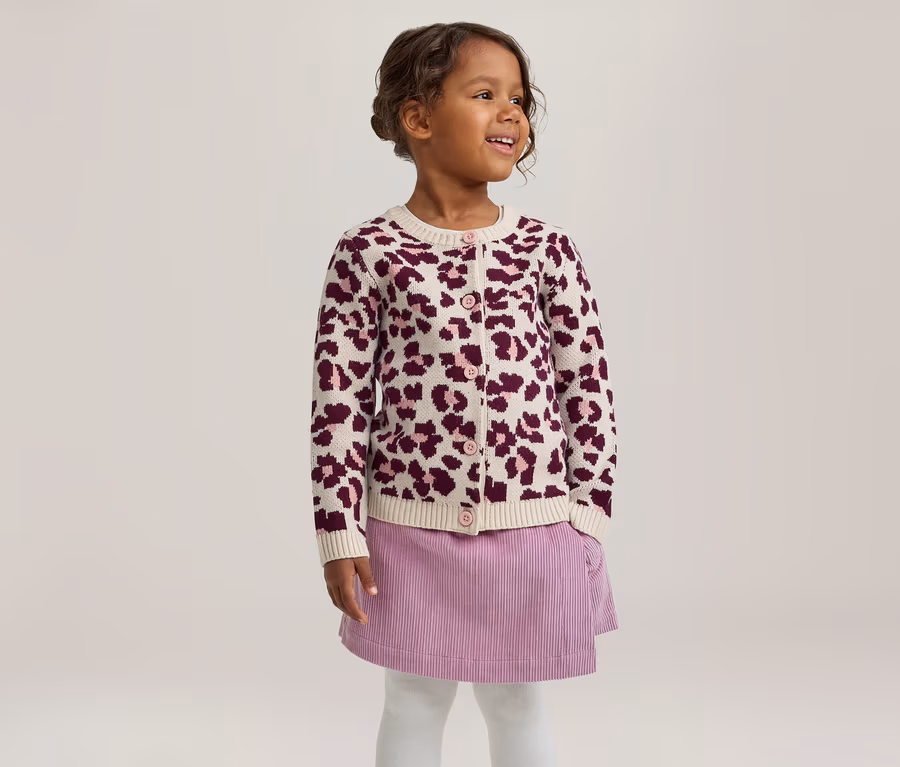 Lächelndes kleines Mädchen, das nach rechts schaut, trägt eine cremefarbene Strickjacke mit lila Leopardenmuster, einen lila Cordrock und weiße Strumpfhosen.