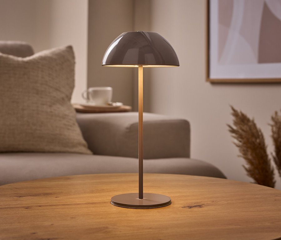 Eine beige LED-Akku-Tischleuchte mit gewölbtem Schirm steht auf einem runden Holztisch. Im Hintergrund sind ein Sofa, eine Tasse und Pampasgras zu sehen.