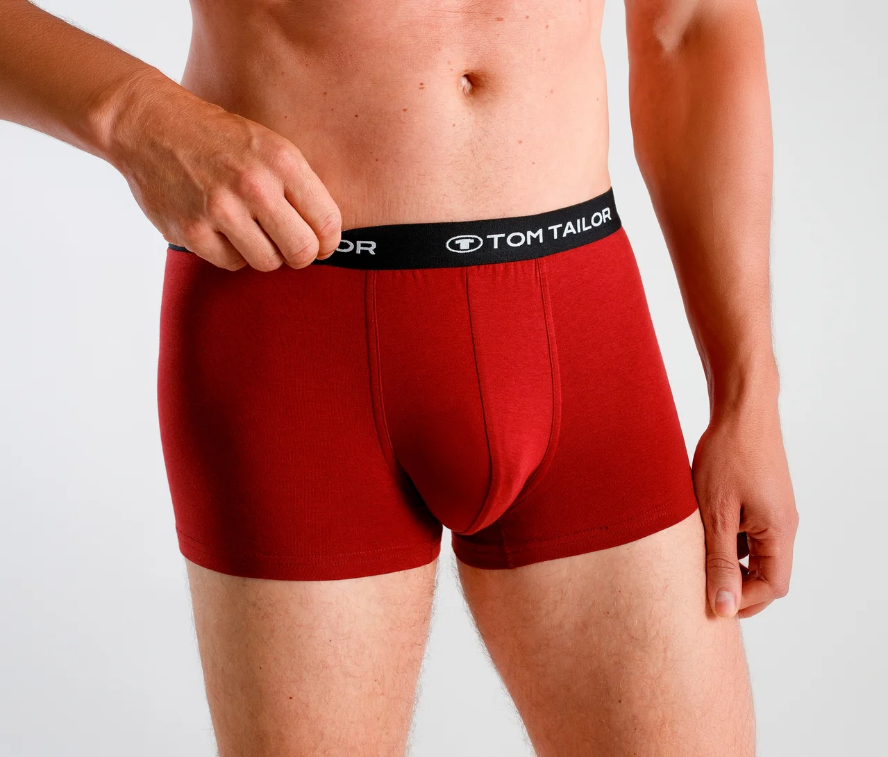 Mann trägt rote Boxershorts mit schwarzem „TOM TAILOR“ Bund und zieht den Bund mit der linken Hand hoch.