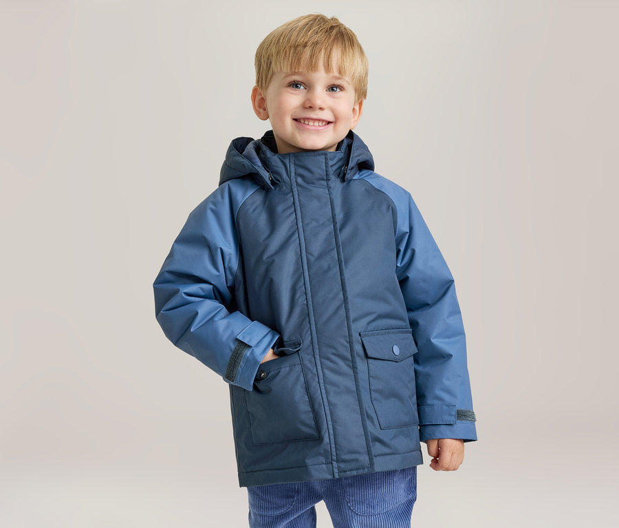 Ein lächelnder kleiner Junge mit blonden Haaren trägt eine zweifarbige blaue Winterjacke, seine linke Hand steckt in der Jackentasche.
