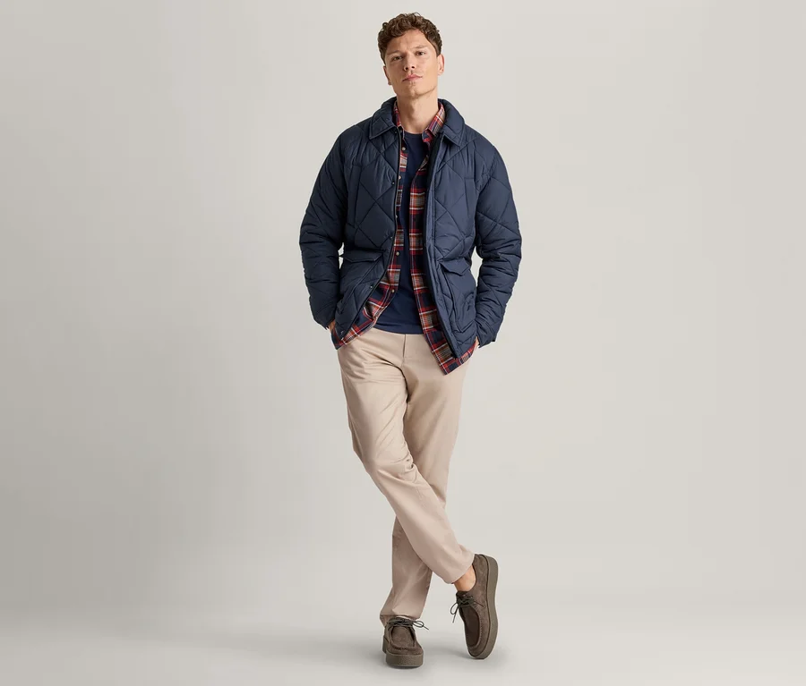 Ein Mann steht mit gekreuzten Beinen und Händen in den Taschen, trägt eine marineblaue Steppjacke über einem rot-blauen Karoshirt, ein dunkelblaues T-Shirt, hellbeige Chinos und dunkelbraune Schnürschuhe.