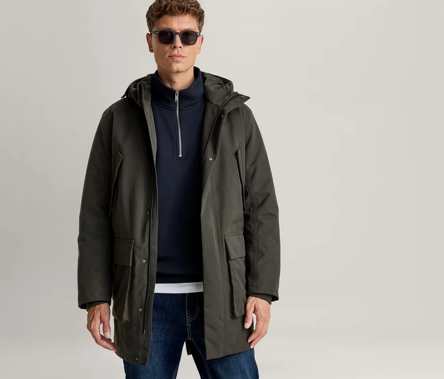 Mann trägt einen dunkelgrünen Kapuzenparka über einem marineblauen Pullover mit Reißverschluss, blaue Jeans und eine Sonnenbrille vor hellem Hintergrund.