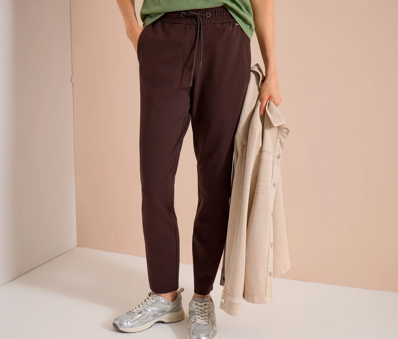 Eine Person trägt eine braune CECIL Jerseyhose, ein grünes T-Shirt und silberne Sneaker, während sie eine beigefarbene Jacke hält.