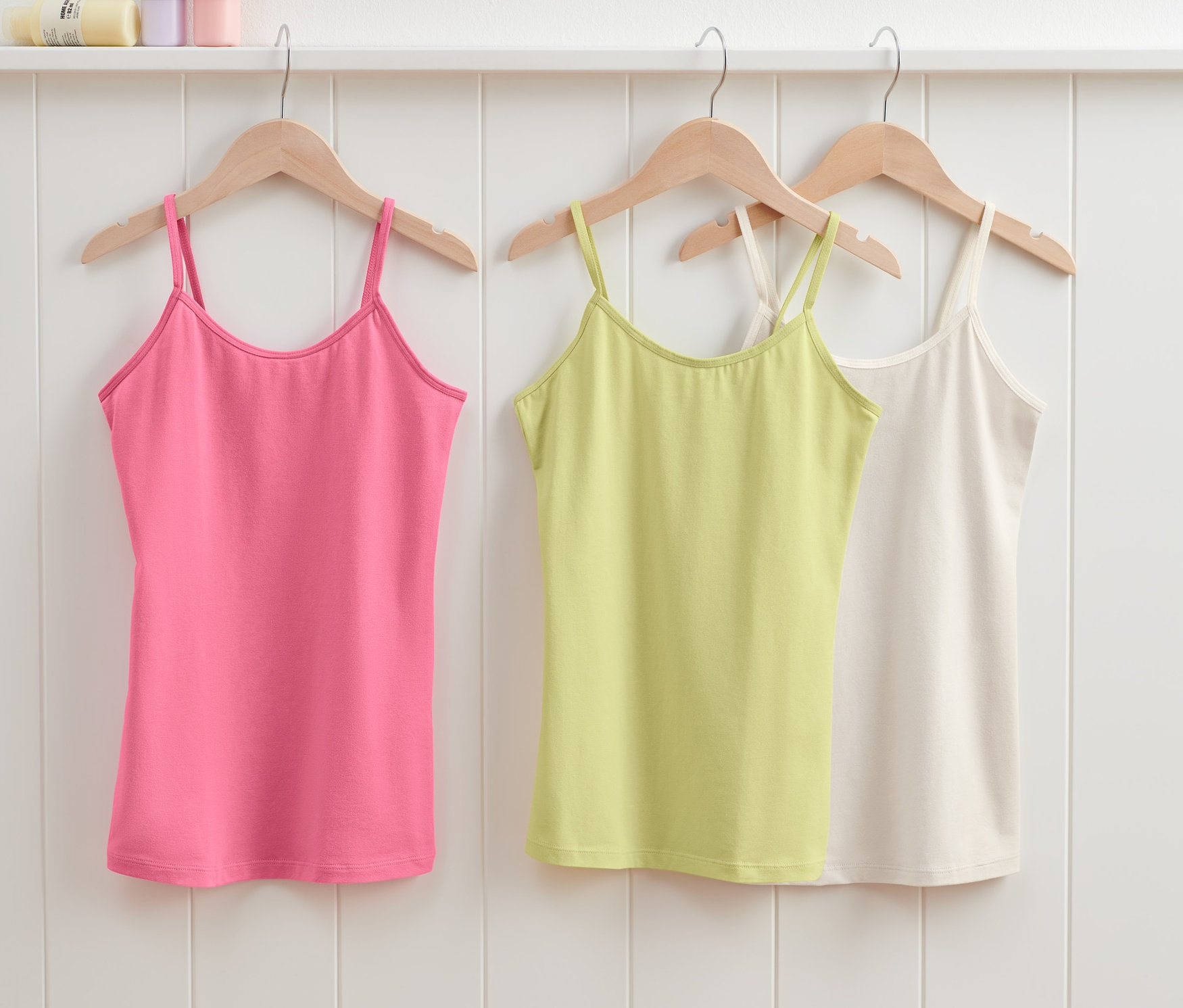 Drei Kinder-Unterhemden (3 Stück) hängen auf Holzkleiderbügeln: rosa, grün und weiß.