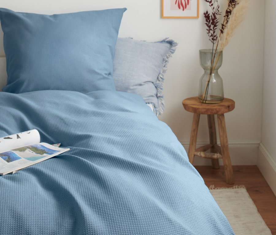 Blaue Waffelpiqué-Bettwäsche in Normalgröße auf einem Bett mit einer Zeitschrift. Neben dem Bett steht ein Holzhocker mit einer Vase und Blumen.