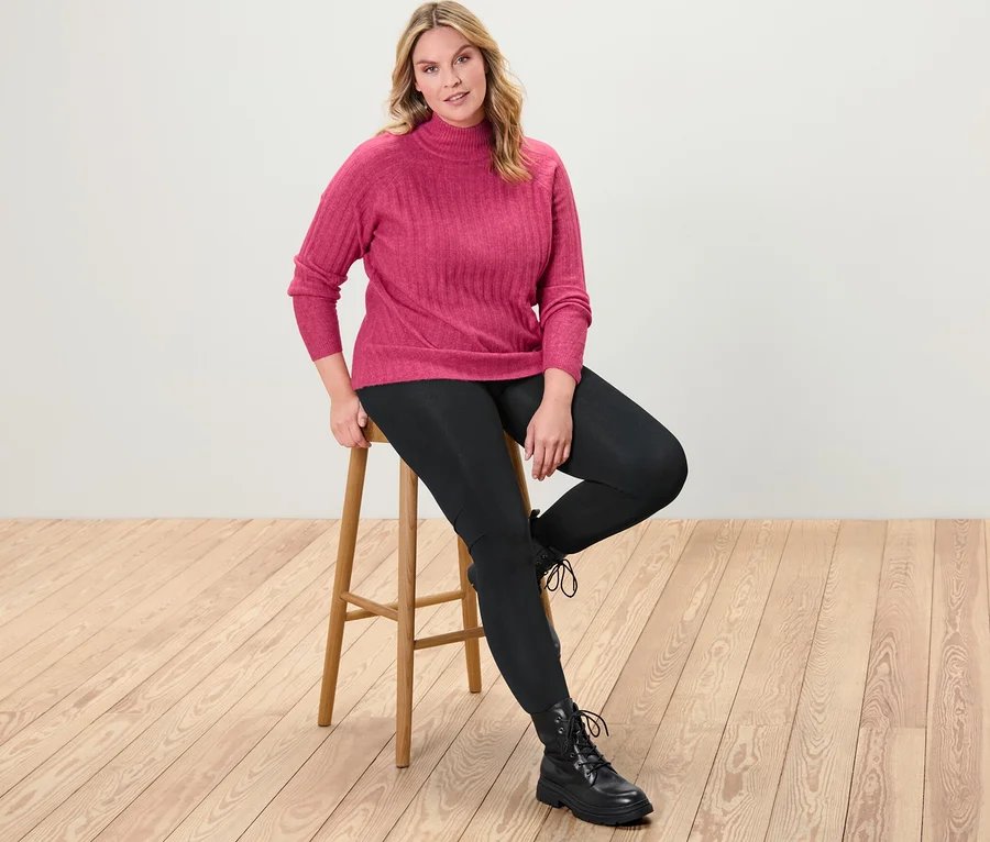 Frau sitzt auf einem Hocker und trägt einen gerippten Strickpullover mit Stehkragen und Thermoleggings.