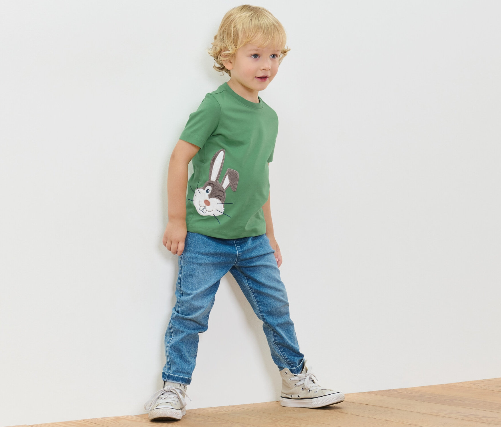 Ein Junge steht an einer Wand und trägt ein grünes T-Shirt mit Applikation und Jeans.