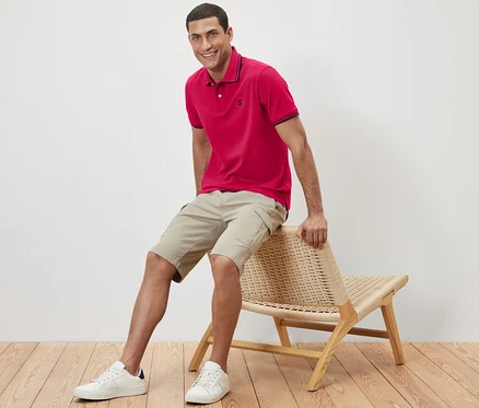 Mann sitzt auf einem Korbstuhl und trägt ein rosa Piqué-Poloshirt.