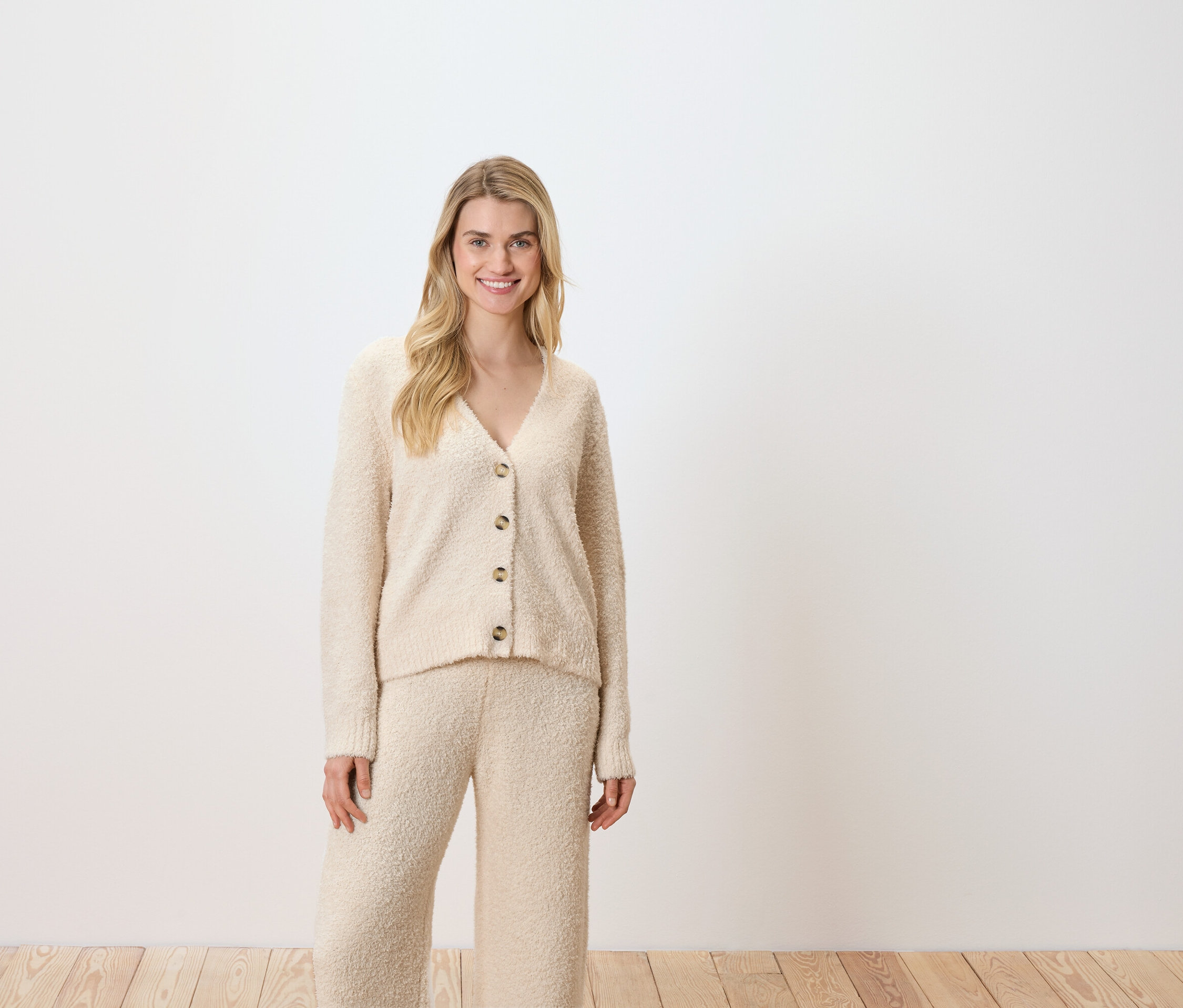 Frau posiert vor einem weißen Hintergrund und trägt eine cremefarbene Strickjacke und Hose.