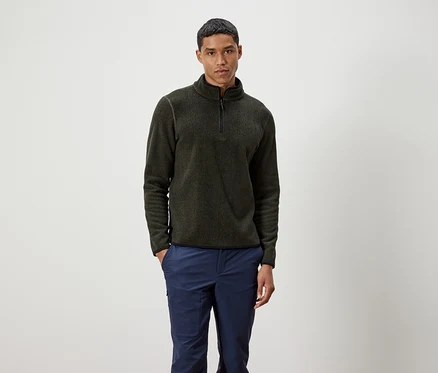 Mann steht und trägt einen olivgrünen Pullover mit Reißverschluss und blaue Hosen.