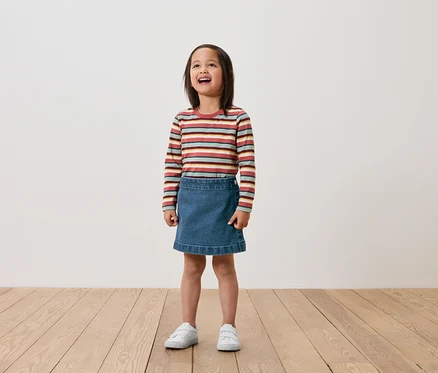Ein Mädchen steht auf einem Holzboden und trägt ein langärmliges Ringelshirt, einen Jeansrock und weiße Turnschuhe.